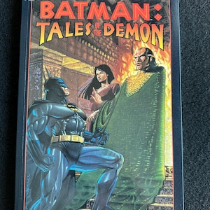 May include: A comic book titled "Batman: Tales of the Demon" featuring Batman, a woman, and a villain in front of a fiery background. The cover has the DC Comics logo and the text "Introduction by Sam Hamm, Afterword by Dennis O'Neil".