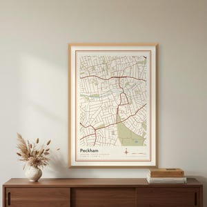 May include: A framed wall art print featuring a detailed street map of Peckham, London, United Kingdom. The map uses a colour palette of cream, sage green, and dark red lines to depict roads and areas, with the text Peckham printed at the bottom.