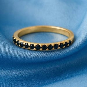 May include: A gold band ring with a row of small, black gemstones. The ring is set against a blue fabric background, creating a contrast between the gold and black of the ring and the blue of the background.