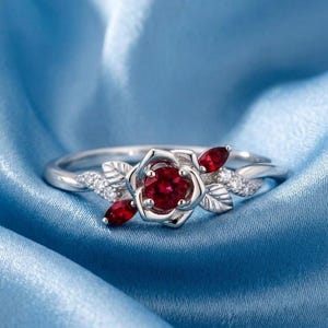 Könnte beinhalten: Ein silberfarbener Ring mit einem zentralen runden roten Edelstein, der von metallenen Blütenblättern in Rosenform umgeben ist. Die Ringschiene ist mit kleinen klaren Steinen und zwei marquise-geschliffenen roten Steinen verziert.