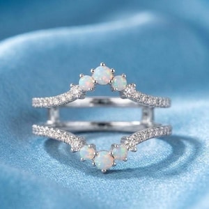 May include: A silver ring guard with a curved design, set with small white opals and sparkling clear stones. The ring is designed to sit around another ring, adding a decorative element. The ring is displayed on a light blue satin surface.
