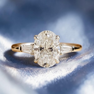 May include: A gold ring with a large oval-cut diamond as the centerpiece, flanked by two smaller, rectangular diamonds. The ring's band is a simple, polished gold. The background is a soft, blue fabric.