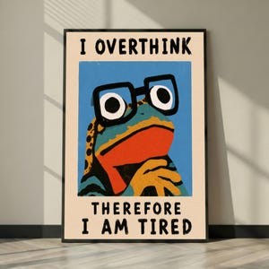 May include: A rectangular wall art print featuring a stylised illustration of a frog wearing thick black glasses against a blue background. The text I OVERTHINK at the top and THEREFORE I AM TIRED at the bottom are printed in black block letters.