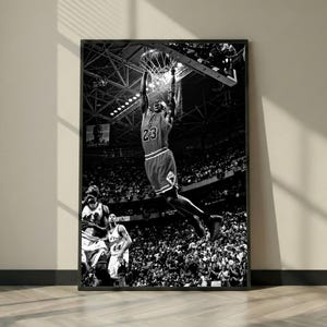 May include: A black and white framed wall art print depicting a basketball player wearing a jersey with the number 23, captured in mid-air while performing a slam dunk into a hoop, set within a crowded arena with stadium lights visible above.