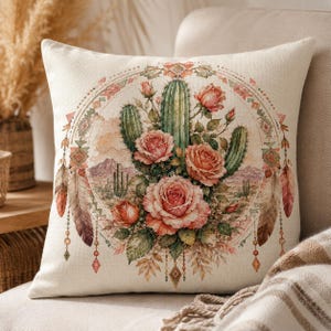 Puede incluir: Un coj&iacute;n cuadrado con funda de tela color crema que presenta un estampado circular central. El dise&ntilde;o muestra cactus verdes y rosas rosadas sobre un paisaje des&eacute;rtico, enmarcado por plumas colgantes y patrones geom&eacute;tricos en tonos suaves.
