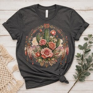 Puede incluir: Una camiseta de manga corta en color gris carb&oacute;n oscuro con un estampado gr&aacute;fico circular. El dise&ntilde;o muestra un paisaje des&eacute;rtico con cactus verdes y rosas rosadas, enmarcado por patrones geom&eacute;tricos y plumas colgantes en tonos tierra.