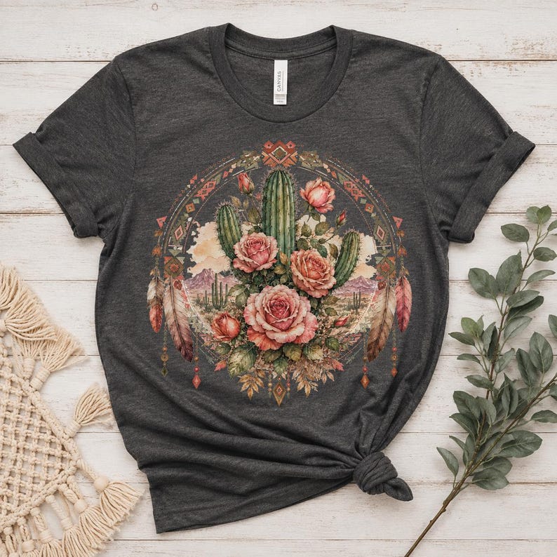 Puede incluir: Una camiseta de manga corta en color gris carb&oacute;n oscuro con un estampado gr&aacute;fico circular. El dise&ntilde;o muestra un paisaje des&eacute;rtico con cactus verdes y rosas rosadas, enmarcado por patrones geom&eacute;tricos y plumas colgantes en tonos tierra.