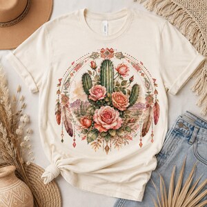 Puede incluir: Una camiseta de manga corta color crema con un estampado gr&aacute;fico circular. El dise&ntilde;o muestra cactus verdes y rosas rosadas centrados en un marco decorado con patrones geom&eacute;tricos, plumas colgantes y un fondo de paisaje des&eacute;rtico.