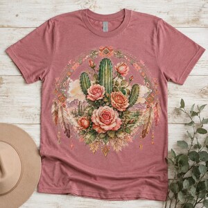 Puede incluir: Una camiseta de manga corta color malva con un gr&aacute;fico central de cactus verdes y rosas rosadas dispuestas en un marco circular. El dise&ntilde;o incluye plumas decorativas y formas geom&eacute;tricas de diamante que rodean la ilustraci&oacute;n floral.