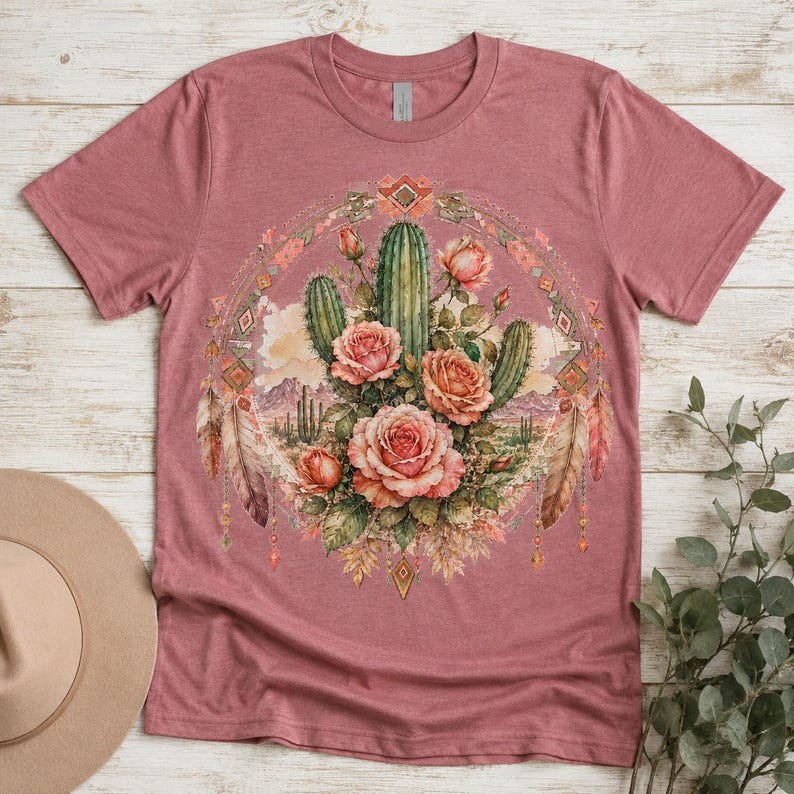 Puede incluir: Una camiseta de manga corta color malva con un gr&aacute;fico central de cactus verdes y rosas rosadas dispuestas en un marco circular. El dise&ntilde;o incluye plumas decorativas y formas geom&eacute;tricas de diamante que rodean la ilustraci&oacute;n floral.