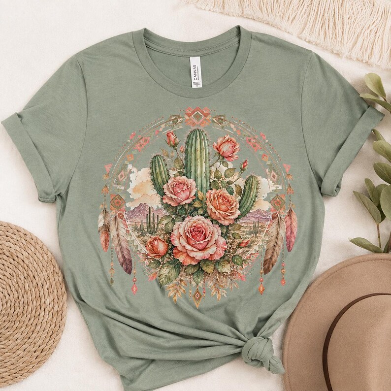 Puede incluir: Una camiseta de manga corta color verde salvia con un estampado gr&aacute;fico central de un paisaje des&eacute;rtico con cactus y rosas rosadas. El dise&ntilde;o est&aacute; enmarcado por un borde circular con patrones geom&eacute;tricos y plumas colgantes en tonos rosa y marr&oacute;n.