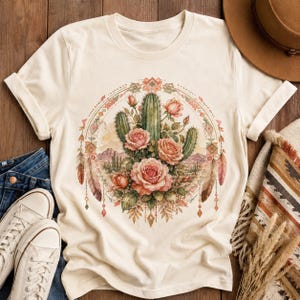 Puede incluir: Una camiseta de manga corta color crema con un estampado gr&aacute;fico circular. El dise&ntilde;o muestra cactus verdes y rosas rosadas centradas dentro de un borde geom&eacute;trico, acentuado por plumas colgantes y un fondo de paisaje des&eacute;rtico.