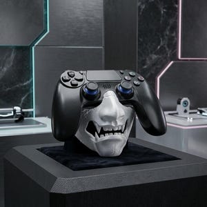 May include: A black PlayStation 4 controller rests atop a white Oni mask, displayed on a black pedestal. The controller has blue accents on the joysticks. The mask features sharp teeth and a detailed design. The background is a modern, dark-toned room.