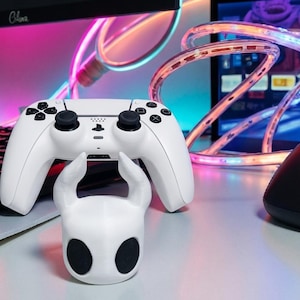 Pode incluir: Um controle branco do PlayStation 5 e uma estatueta branca de um personagem com grandes olhos negros e chifres. O controle tem botões pretos e pads direcionais. Um mouse preto e um monitor de computador com luzes coloridas estão ao fundo.