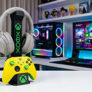 Op de afbeelding: Een gaming-opstelling met een zwart-gele Xbox-controllerstandaard met een headset en controller. De standaard heeft het Xbox-logo. Een gaming-pc en monitor staan op de achtergrond.