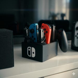 Pode incluir: Base de carregamento preta com o logotipo branco da Nintendo, com comandos Joy-Con azuis e vermelhos. Cabos e um comando preto também são visíveis. A base está sobre uma superfície branca.