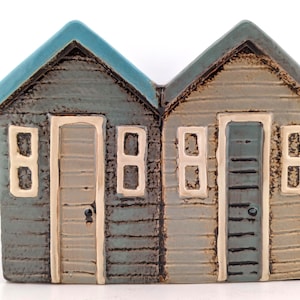 May include: A ceramic ornament depicting two adjoining miniature houses. The houses have blue roofs, light blue and tan siding, and cream-coloured trim around the doors and windows. Each house has a door and two windows. The piece is on a white background.