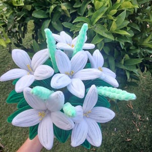 Peut inclure: Un bouquet de fleurs de lys blanches fabriquées à la main à partir de cure-pipes duveteux. Les fleurs présentent des centres jaunes et sont agrémentées de tiges vert clair duveteuses et de feuilles vert foncé, créant un arrangement floral texturé.