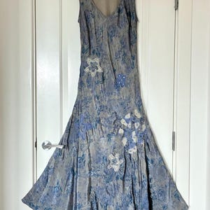 May include: A sleeveless, V-neck midi dress in a muted grey-blue tone featuring an all-over floral pattern. The garment is accented with several white, star-shaped fabric appliqués and a cluster of white floral embroidery on the lower right side.