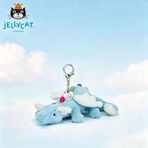 Puede incluir: Un llavero de dragón de peluche azul claro con alas y cuernos blancos. Tiene un llavero plateado y una pequeña etiqueta. El logotipo de Jellycat es visible sobre un fondo de cielo azul. El dragón tiene acentos coloridos.