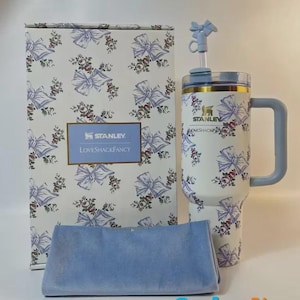 May include: A white Stanley tumbler with a floral and bow pattern, a blue handle, and a matching box. The box and tumbler feature the "LoveShackFancy" logo. A blue cloth is in front of the box.