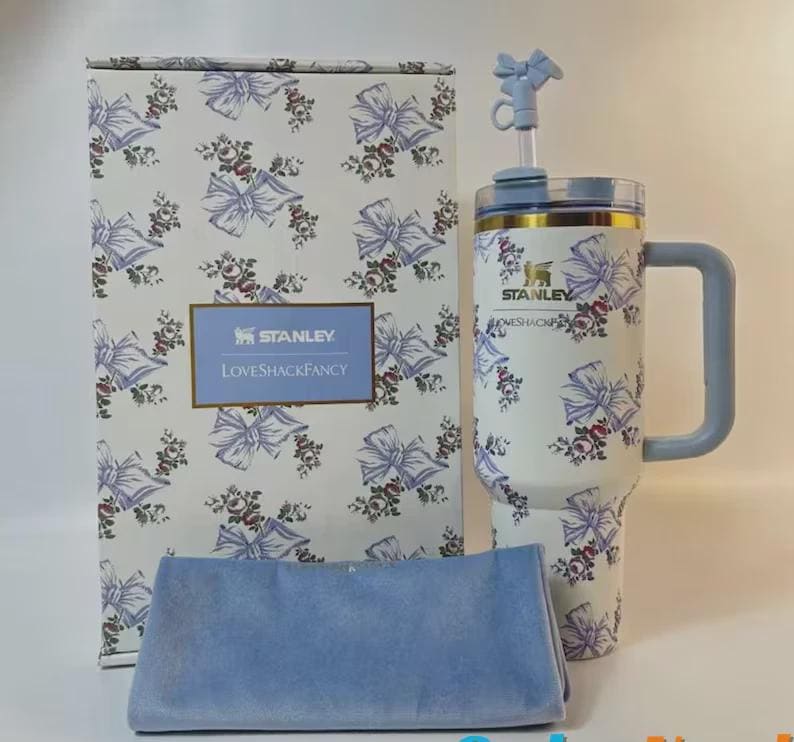 May include: A white Stanley tumbler with a floral and bow pattern, a blue handle, and a matching box. The box and tumbler feature the "LoveShackFancy" logo. A blue cloth is in front of the box.
