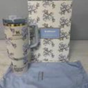 May include: A white travel mug with a blue floral pattern, a handle, and a clear lid. The mug is next to a matching box and a blue cloth. The mug has the word "Stanley" printed on it.