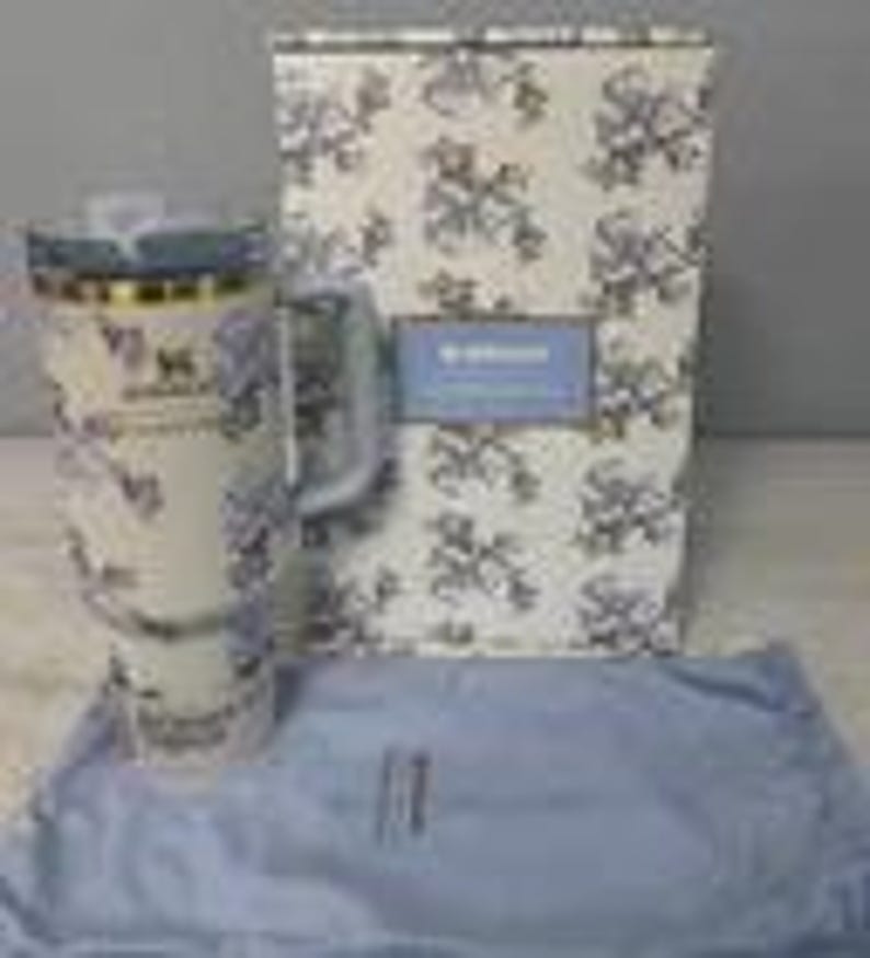 May include: A white travel mug with a floral pattern and a handle, accompanied by a matching box and a blue cloth. The mug has a clear lid and gold accents. The box features a similar floral design.