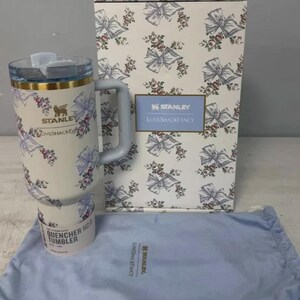 May include: A white Stanley Quencher H2.0 tumbler with a floral and bow pattern, a handle, and a clear lid. The tumbler is packaged with a matching box and a blue drawstring bag. The box features the Stanley and LoveShackFancy logos.