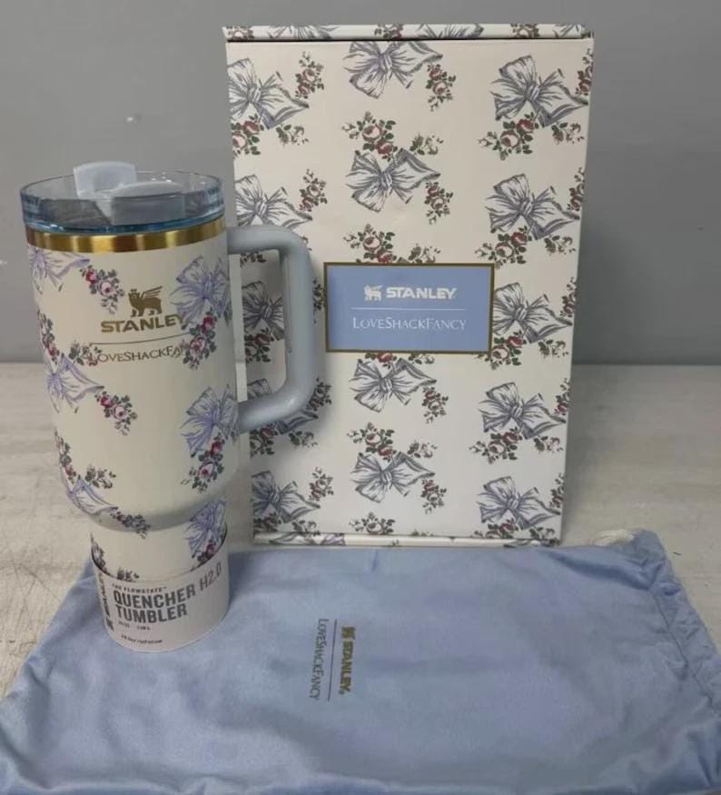 May include: A white Stanley Quencher H2.0 tumbler with a floral and bow pattern, a handle, and a clear lid. The tumbler is packaged with a matching box and a blue drawstring bag. The box features the Stanley and LoveShackFancy logos.
