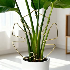 May include: A gold-coloured metal plant support in the shape of a stylized leaf, placed in a white ceramic pot with a green plant. The support has a geometric design with curved and angular lines, providing a decorative and functional element.