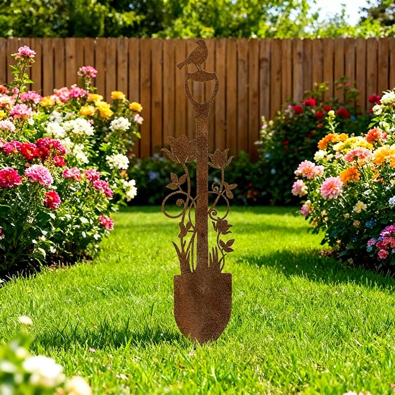 Peut inclure: Sculpture d&eacute;corative de pelle de jardin en m&eacute;tal rouill&eacute;, avec un oiseau perch&eacute; sur la poign&eacute;e. La pelle pr&eacute;sente des d&eacute;coupes florales et de feuilles. La sculpture est plac&eacute;e dans un jardin avec des fleurs color&eacute;es et une cl&ocirc;ture en bois en arri&egrave;re-plan.