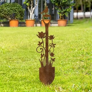 Peut inclure: Une pelle de jardin d&eacute;corative en m&eacute;tal avec un oiseau perch&eacute; sur la poign&eacute;e. La pelle pr&eacute;sente un motif floral avec des feuilles et des fleurs. La pelle est en m&eacute;tal brun rouill&eacute; et est plac&eacute;e dans une zone herbeuse.