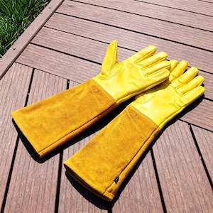 May include: A pair of yellow leather gardening gloves with long, mustard-colored suede sleeves. The gloves are laid out on a wooden surface, showcasing their bright color and protective design. The gloves are designed to protect the hands and arms while gardening.