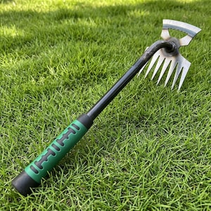 Peut inclure: Un outil de jardinage à main avec un manche noir et une poignée verte. L'outil a une lame métallique d'un côté et des dents de l'autre. L'outil est conçu pour le jardinage et se détache sur un fond d'herbe verte.