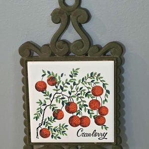 Puede incluir: Salvamanteles de baldosas de arándanos vintage con marco de metal decorativo. La baldosa presenta un diseño de rama de arándanos con bayas rojas y hojas verdes. La palabra "Cranberry" está escrita en cursiva.