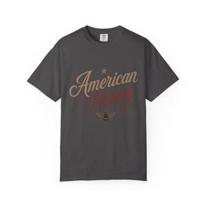 May include: Charcoal grey t-shirt featuring the text "American Honey" in a retro design. The words are in tan and red, with a star and bee illustration. A comfortable tee for casual wear.