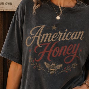 May include: Dark grey t-shirt featuring the text "American Honey" in a vintage style. The design includes a star, a bee, and floral accents. The shirt has short sleeves and a relaxed fit.