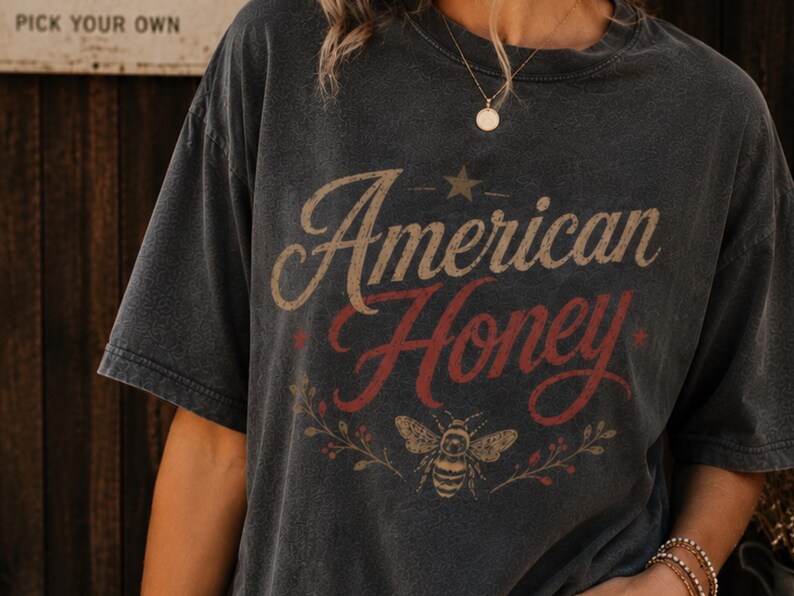 May include: Dark grey t-shirt featuring the text "American Honey" in a vintage style. The design includes a star, a bee, and floral accents. The shirt has short sleeves and a relaxed fit.