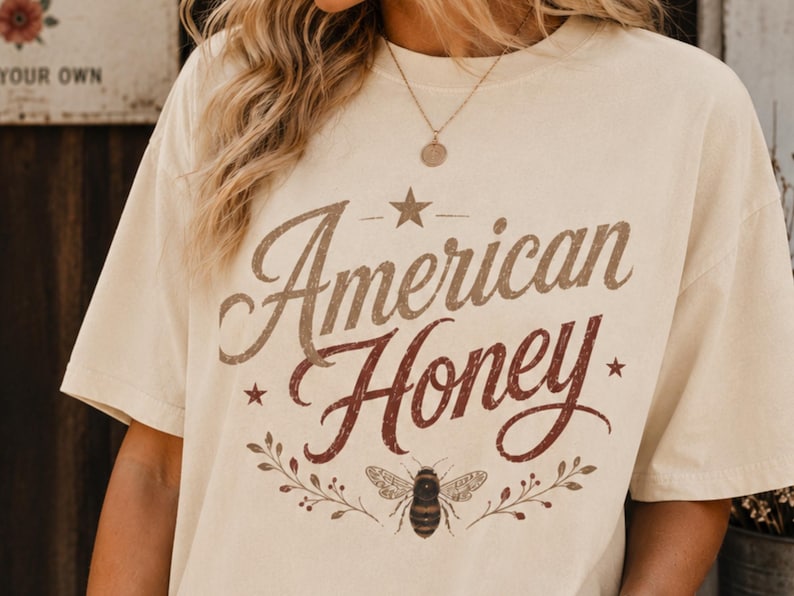 May include: A cream-coloured t-shirt featuring the words "American Honey" in brown and burgundy script. The design includes a star, a bee, and floral elements. The shirt has short sleeves.