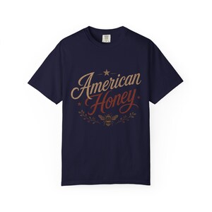 May include: A navy blue t-shirt featuring the text "American Honey" in a vintage style. The design includes a star and a bee graphic, with the text in tan and red. The shirt has a crew neck.