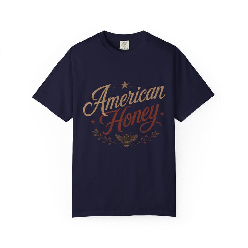 May include: A navy blue t-shirt featuring the text "American Honey" in a vintage style. The design includes a star and a bee graphic, with the text in tan and red. The shirt has a crew neck.