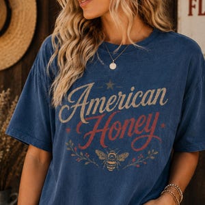 May include: A navy blue t-shirt featuring the text "American Honey" in a vintage style. The design includes a star, a bee, and floral elements. The shirt has a relaxed fit.