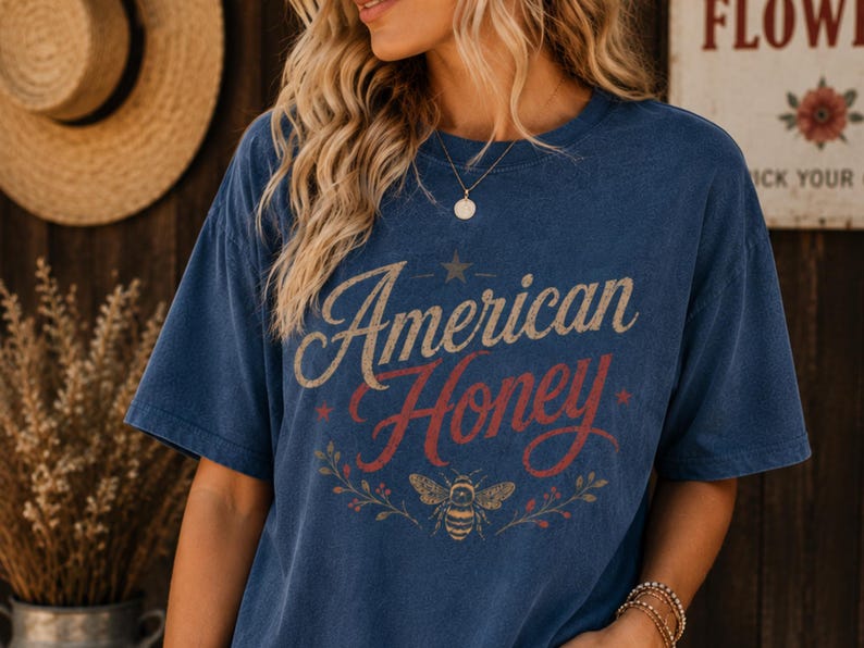 May include: A navy blue t-shirt featuring the text "American Honey" in a vintage style. The design includes a star, a bee, and floral elements. The shirt has a relaxed fit.