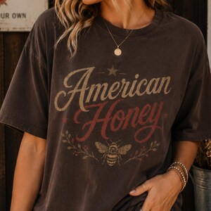 May include: Dark brown t-shirt featuring the text "American Honey" in a vintage style. The design includes a star, a bee, and floral elements. The shirt has short sleeves and a relaxed fit.
