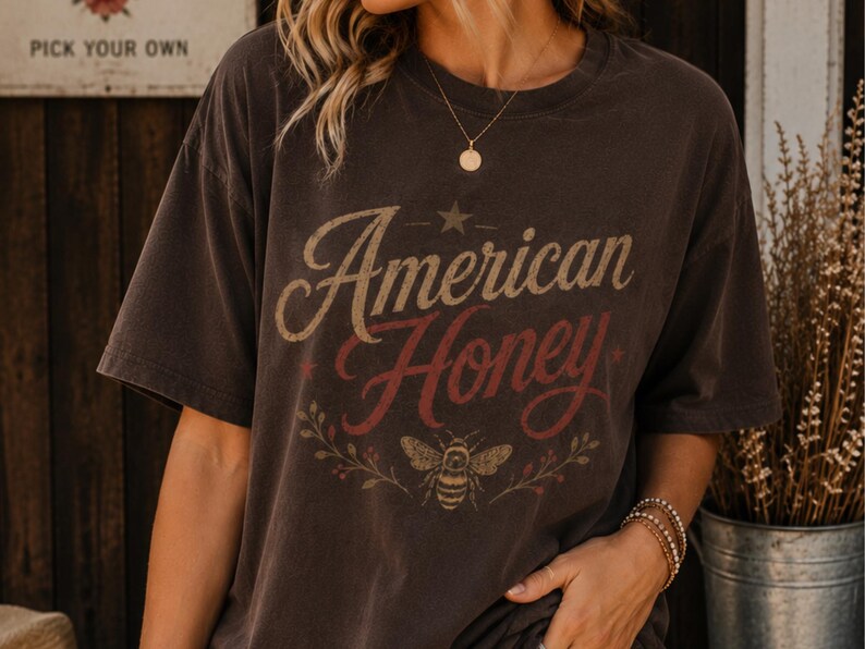 May include: Dark brown t-shirt featuring the text "American Honey" in a vintage style. The design includes a star, a bee, and floral elements. The shirt has short sleeves and a relaxed fit.