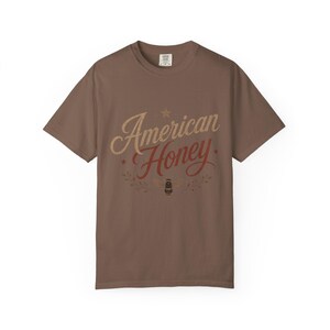 May include: A brown t-shirt featuring the text "American Honey" in a vintage font. The design includes a star, leaves, and a bee illustration. The shirt is a solid brown colour.