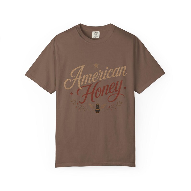 May include: A brown t-shirt featuring the text "American Honey" in a vintage font. The design includes a star, leaves, and a bee illustration. The shirt is a solid brown colour.