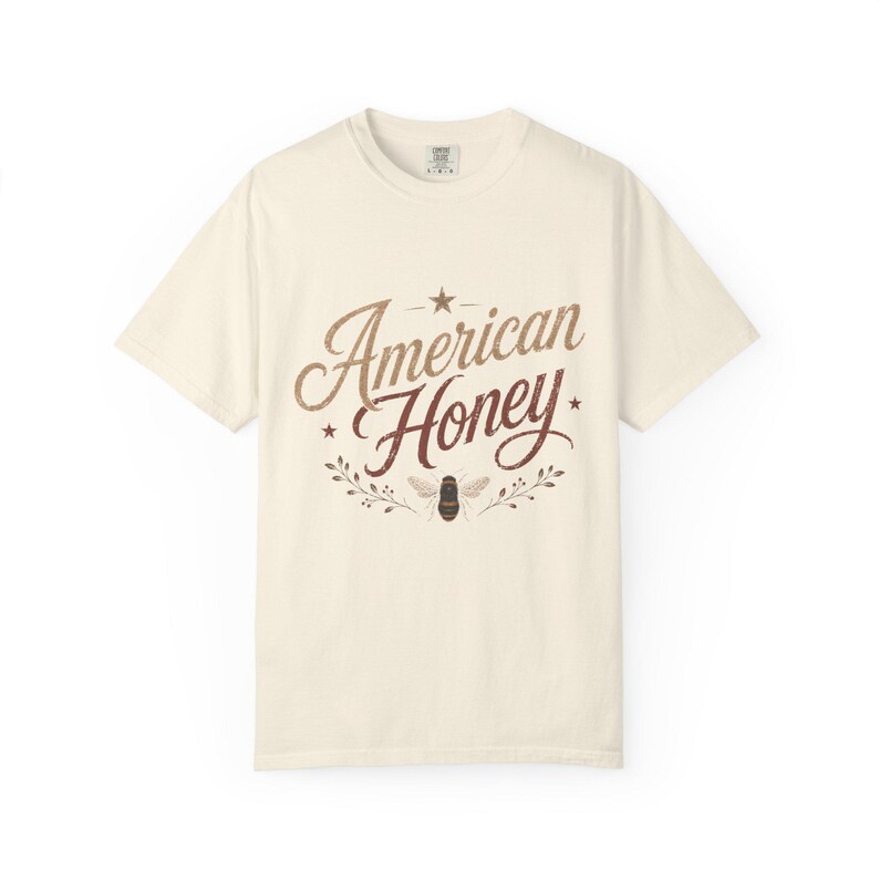 May include: Off-white t-shirt featuring the text "American Honey" in a brown, vintage font. The design includes a star, a bee, and floral elements. The shirt is made of a soft material.