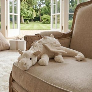 Puede incluir: Un juguete de peluche con forma de dragón de color crema, con alas y una larga cola, descansa sobre un sillón beige. El dragón tiene una expresión amigable y está hecho de un material suave. La silla está en una habitación con una ventana que da a un jardín verde.